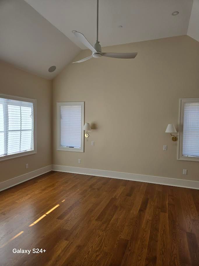 An empty room with light beige walls, hardwood floors, two windows with blinds, wall sconces, and a white ceiling fan.