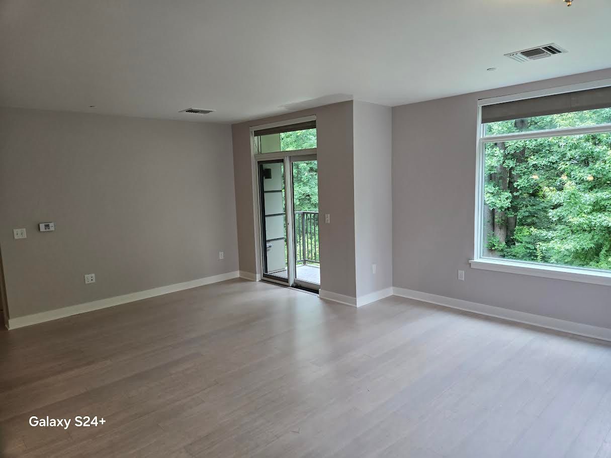 A spacious, empty room with light wood-style flooring, pale gray walls, and a large window overlooking green trees.
