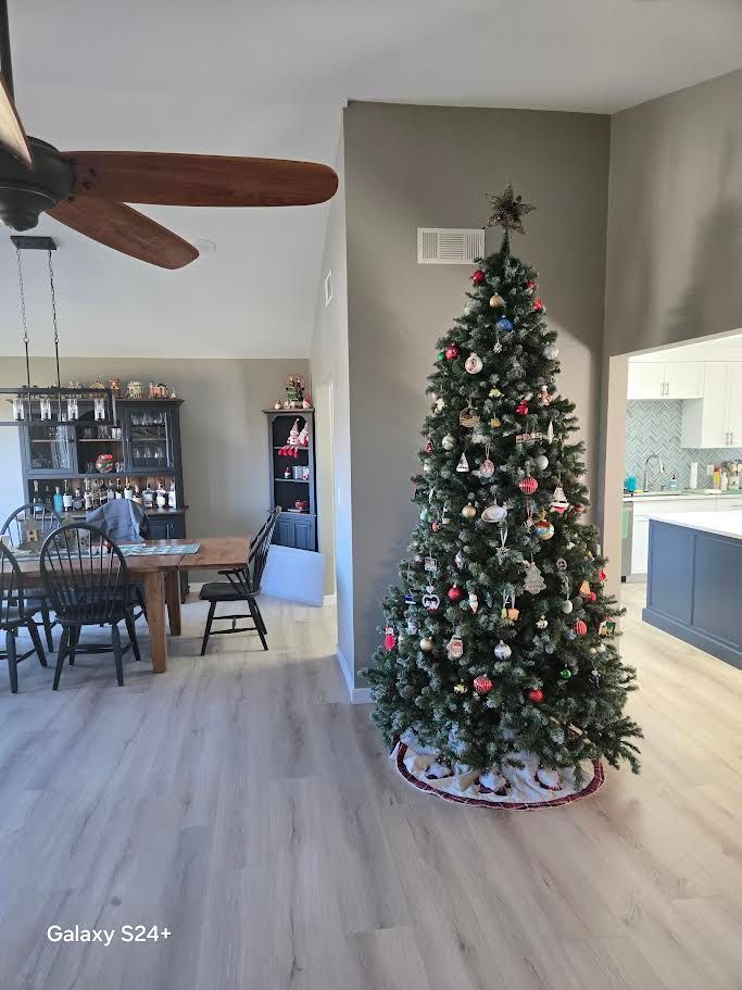 A decorated Christmas tree sits in a dining room with wood-look floors, a wooden dining table, and a dark hutch.