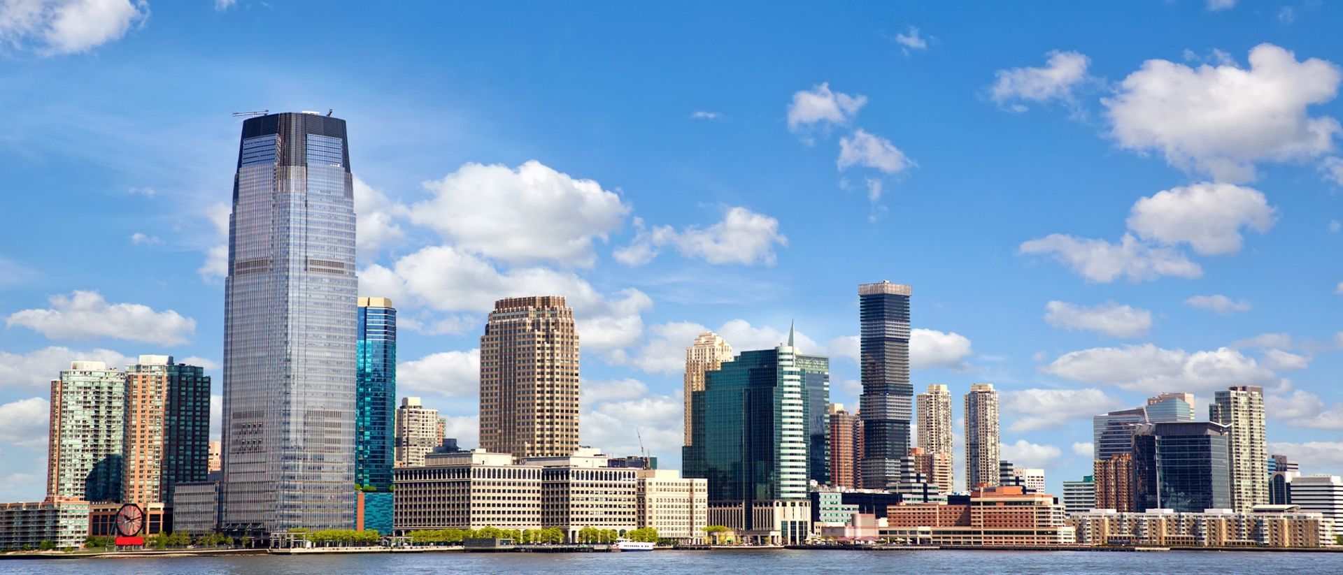 Skyline of Jersey City, NJ, with a cluster of skyscrapers on a sunny day with clouds.