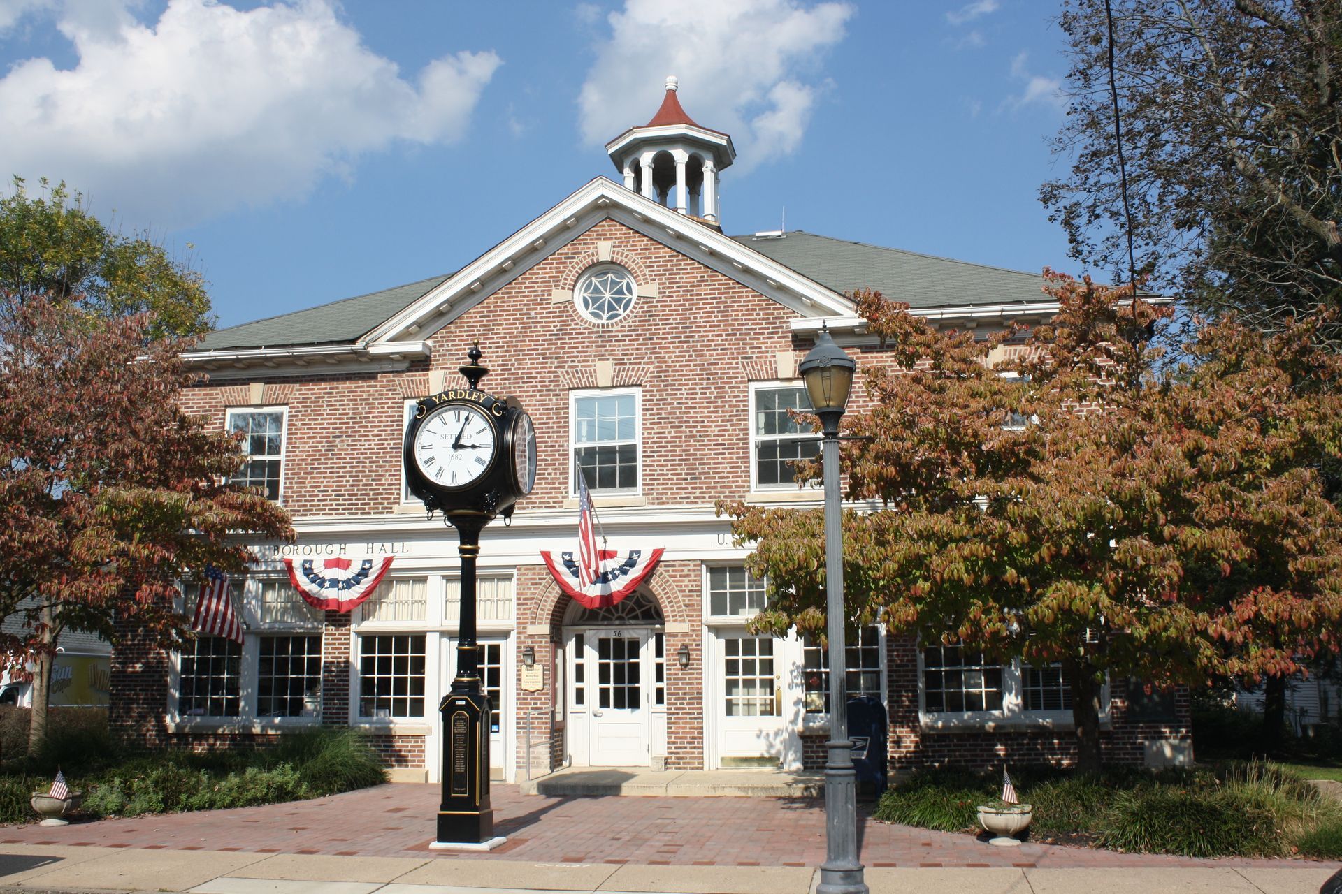Brick building with clock, US flags, trees, and streetlights under a blue sky.