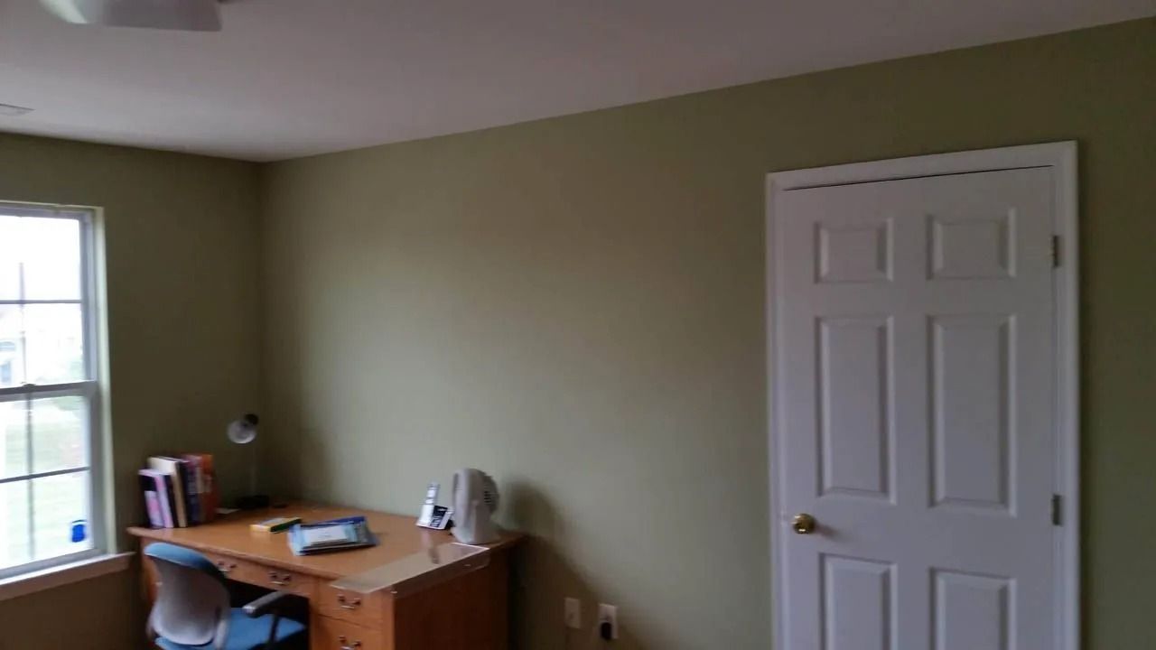 Room with green walls, a white door, a desk with books and a window on the left.