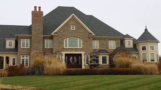 Large brick house with a gray roof and lush green lawn. Tall brown bushes border the house.