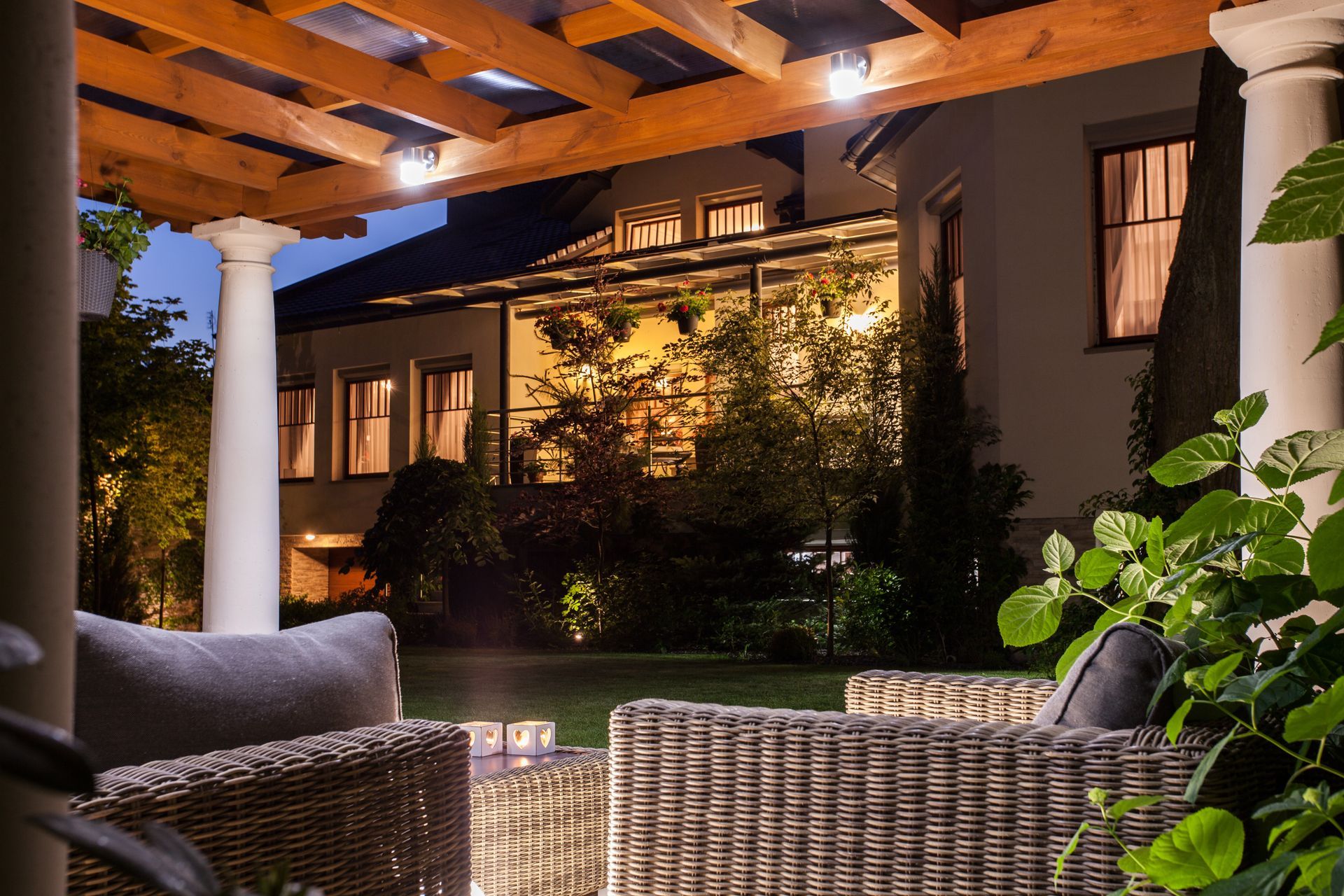 Cozy wicker patio seating under a wooden pergola at dusk, overlooking a lit garden courtyard