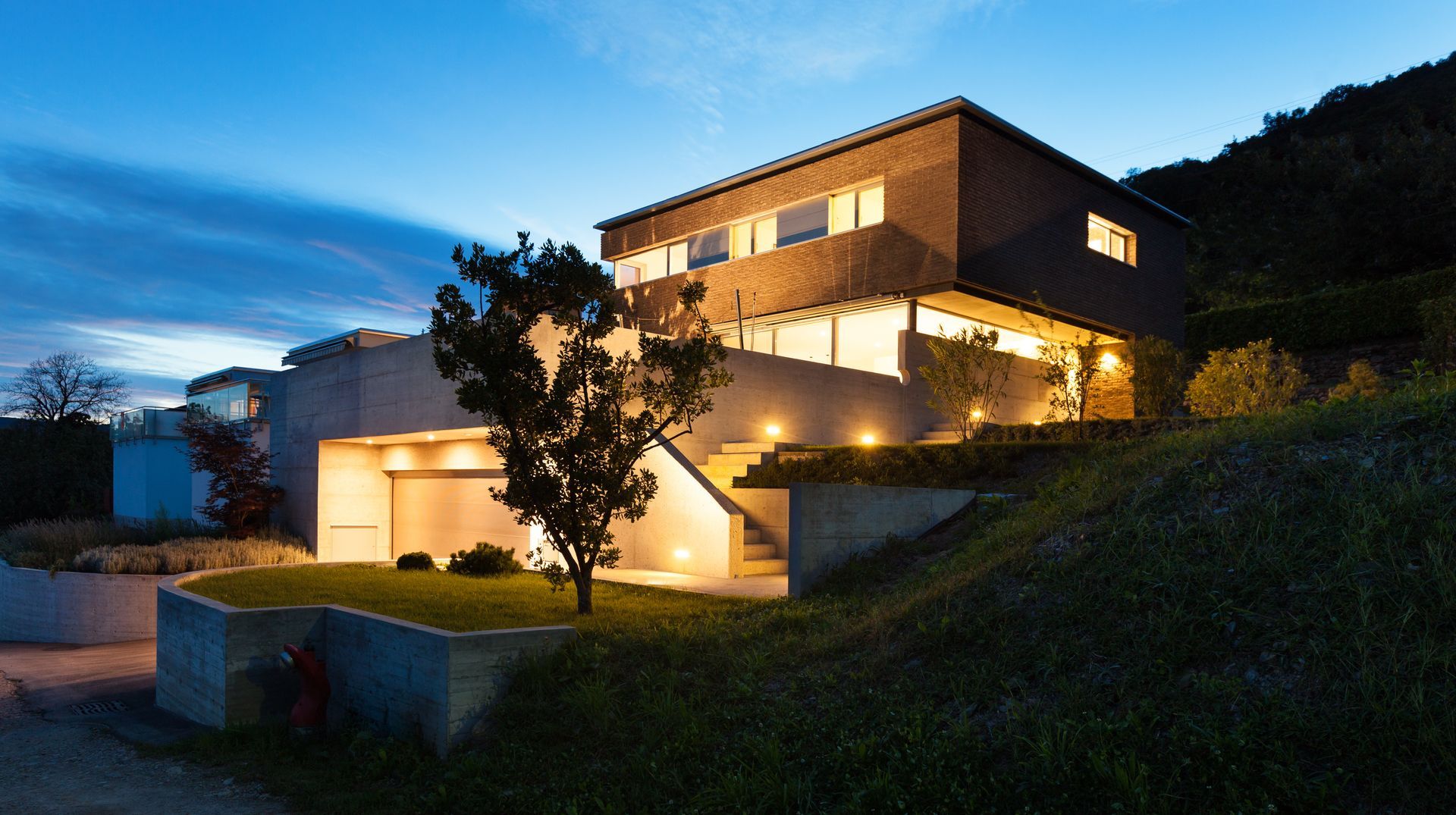 Modern house at dusk with warm exterior lights on a hillside driveway and a lone tree in front