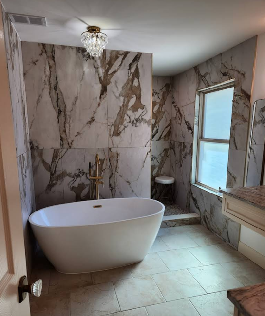 A freestanding white bathtub set against a wall with large, gold-veined white marble tiles and a crystal chandelier above.