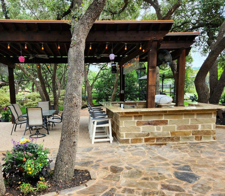 An outdoor patio area with a stone bar, wooden pergola, string lights, and a dining set under leafy trees.
