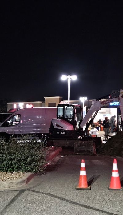 A backhoe next to a building at night, with a white van nearby and traffic cones in the foreground.
