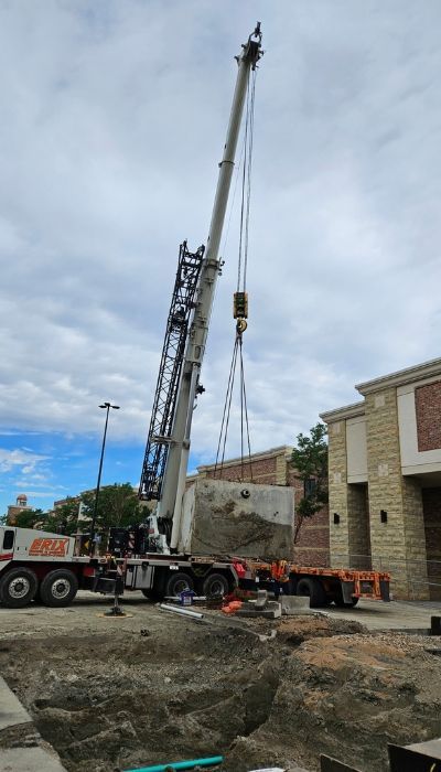 Crane lifting a large concrete block near a building under a cloudy sky.