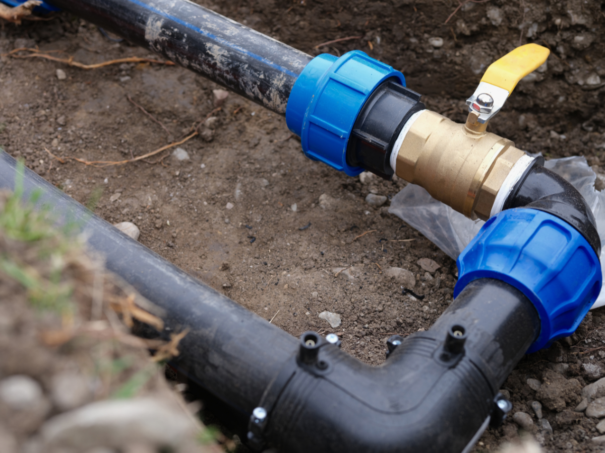 Black water pipes with blue couplings and a brass valve in a dirt trench.