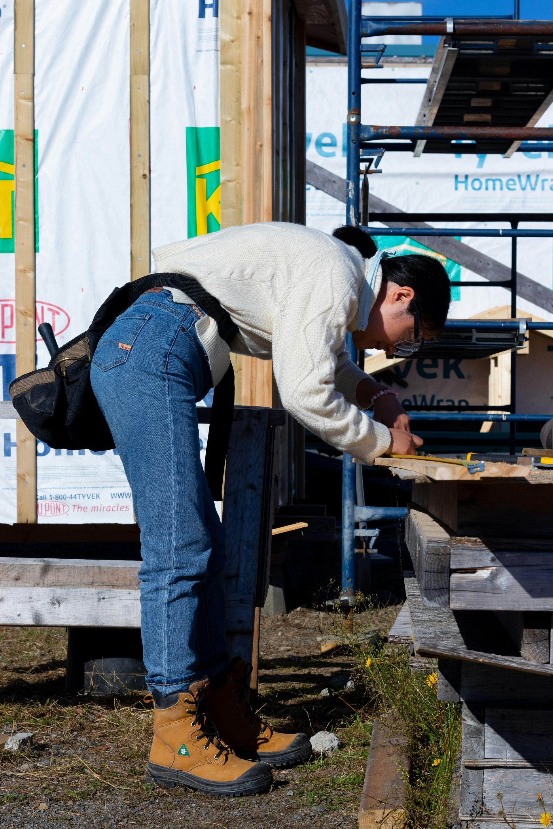 Person in work boots and jeans bending over wood, using tools at a construction site.