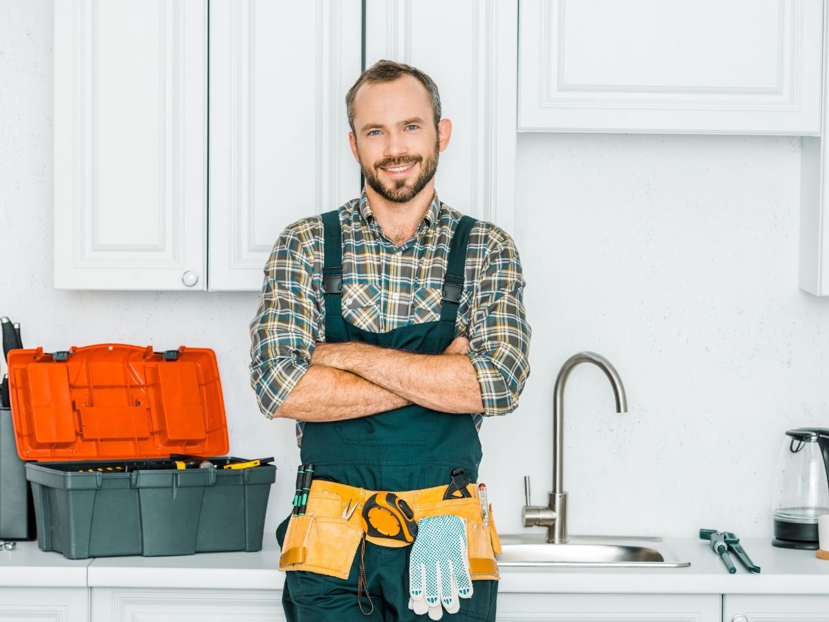 A plumber working under a kitchen sink, using a drain cleaning machine, with an open toolbox nearby.