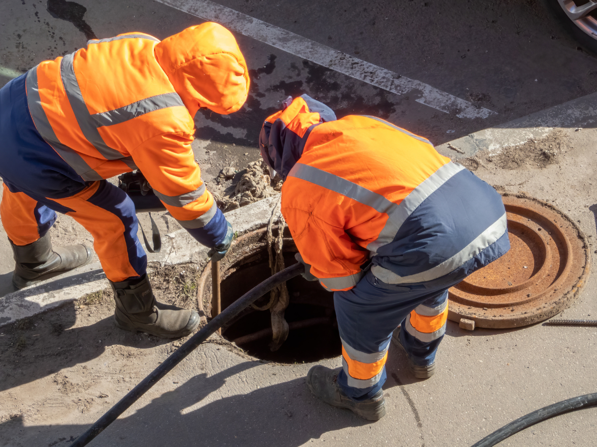Two workers in orange and blue protective gear examine a sewer manhole on a city street.