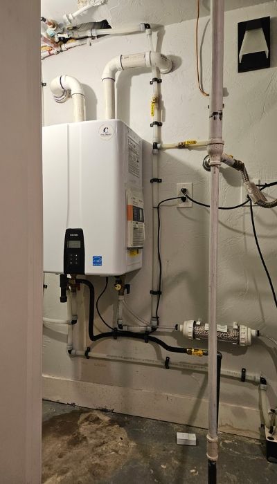 White tankless water heater on a basement wall with pipes, electrical, and a water filter.