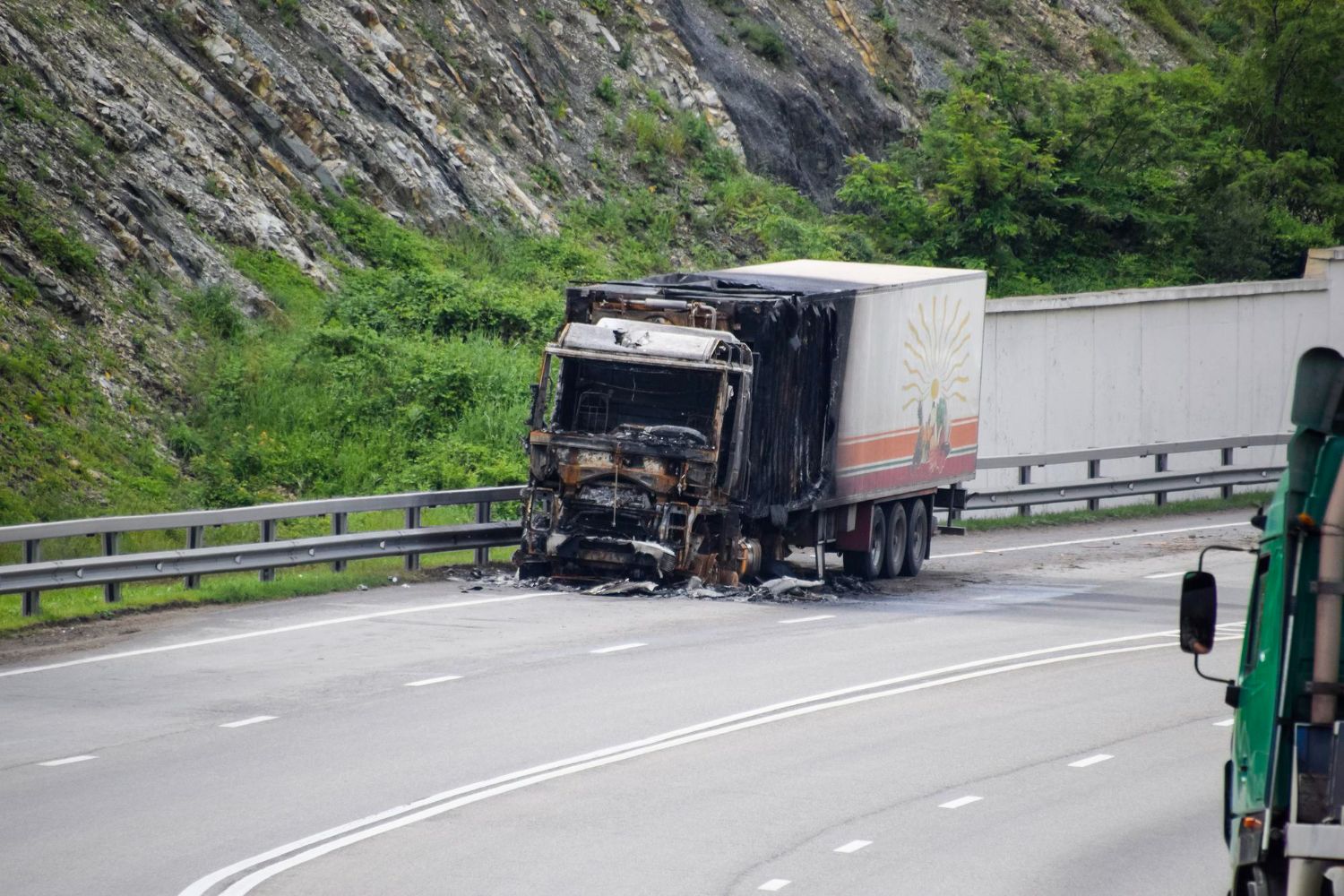 Burned semi-truck on a highway. Blackened cab, white trailer, road, and rocky hillside visible.