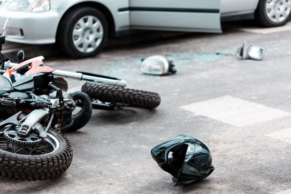 Motorcycle and car collision scene; motorcycle damaged on ground, helmet, broken glass, open car door.