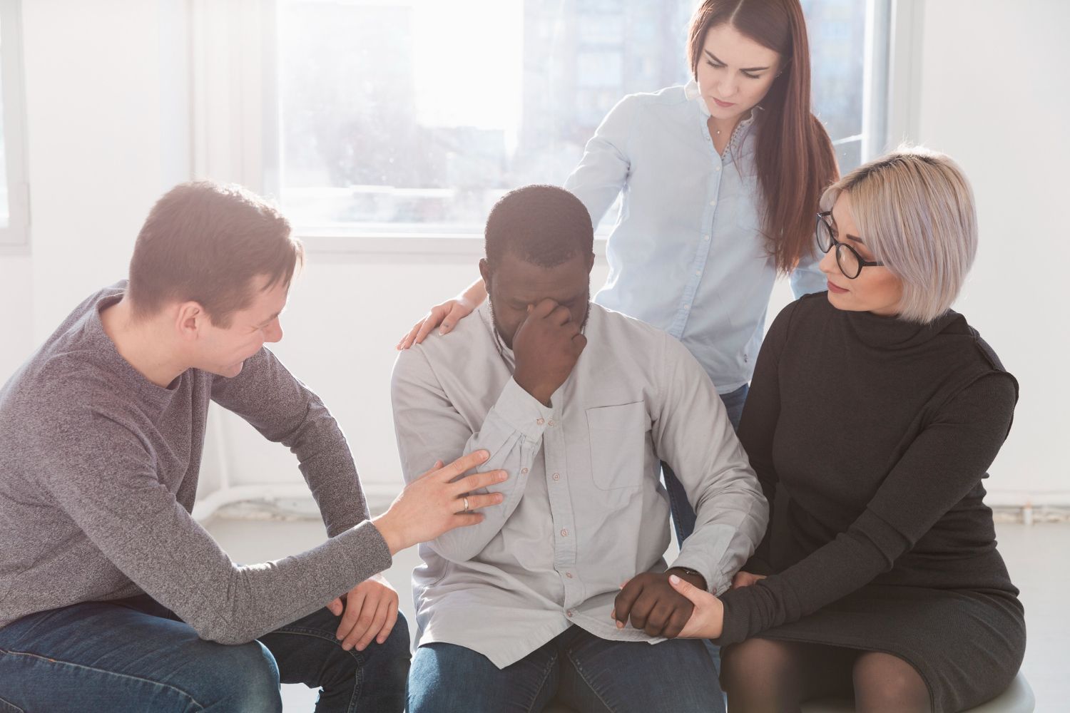 Group supports a Black man, visibly upset, with hands on his shoulders and arms, indoors.