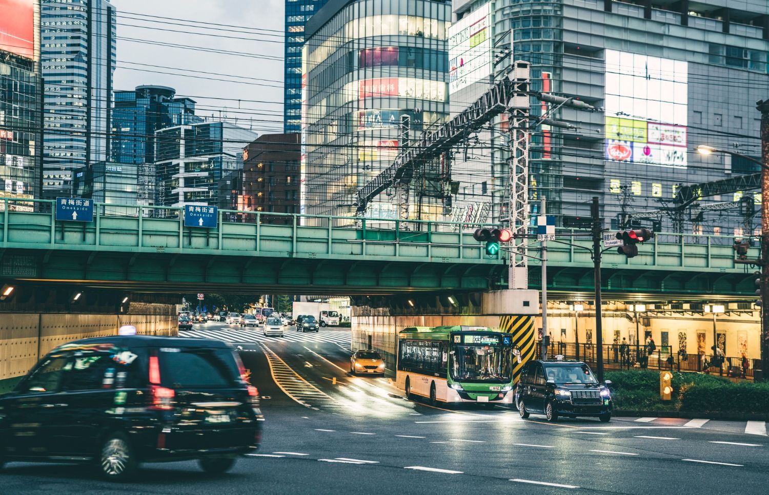 Busy Tokyo street scene with cars, bus, and elevated train tracks. Brightly lit buildings.