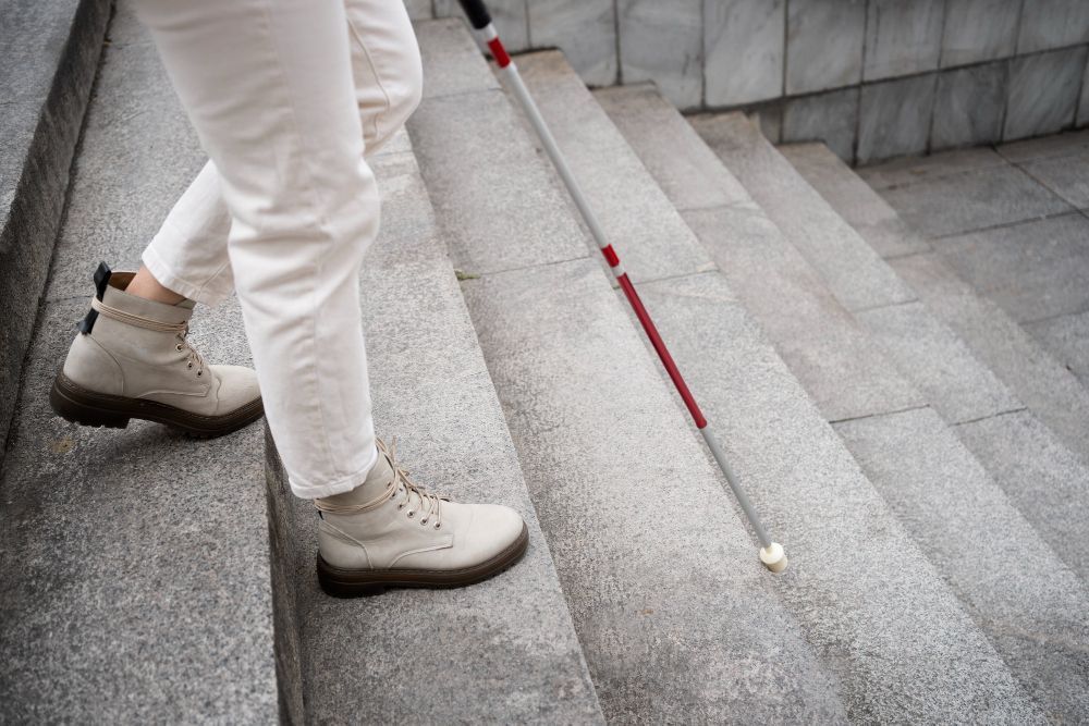Person with a white cane descends stone steps. Beige boots, white pants, and red stripe on the cane.