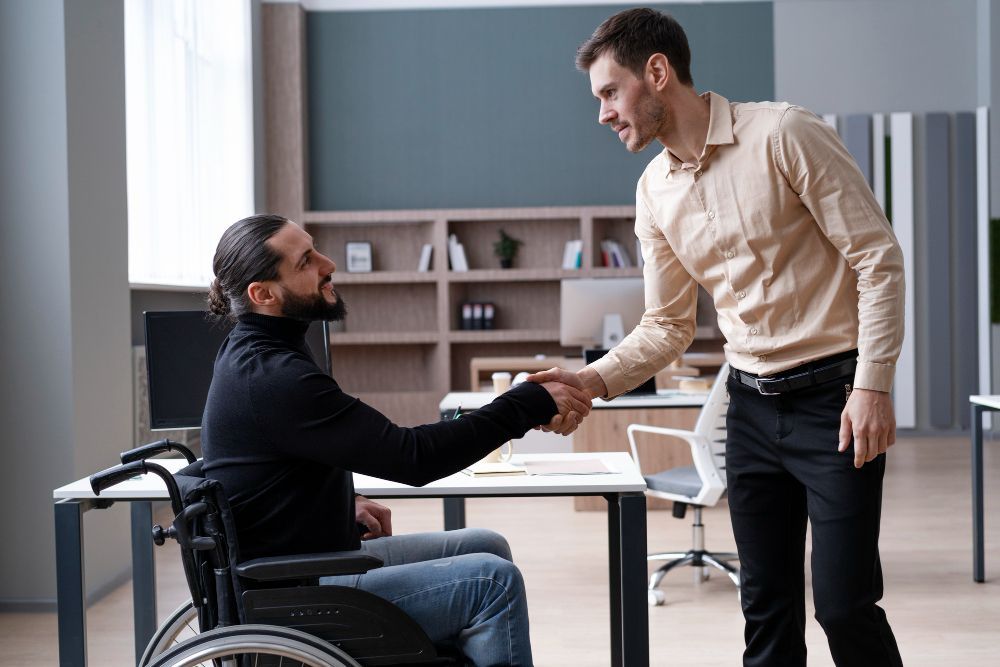Two men shaking hands in an office setting; one in a wheelchair.