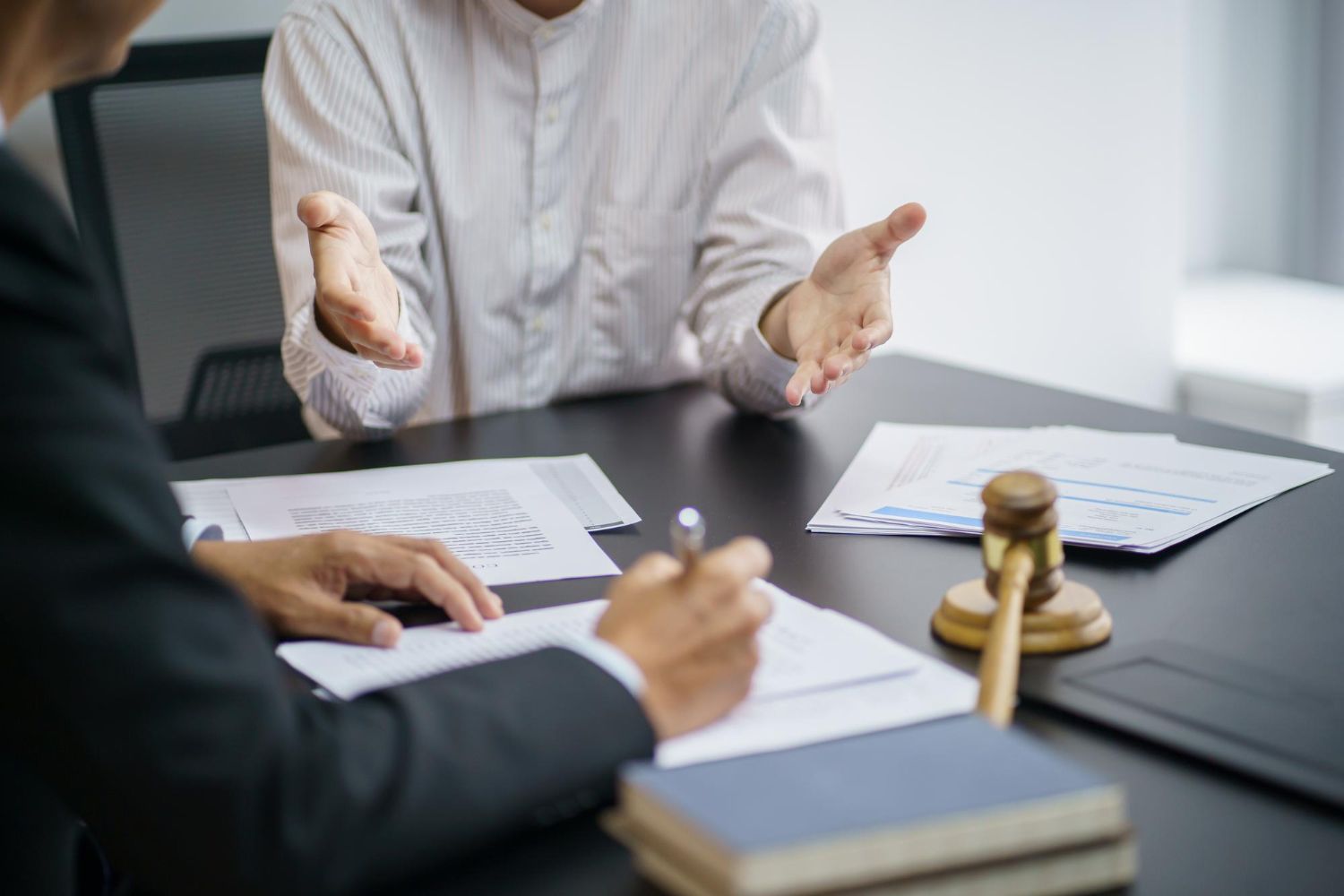 Two people at a table, one gesturing, the other writing. Legal documents and a gavel present.