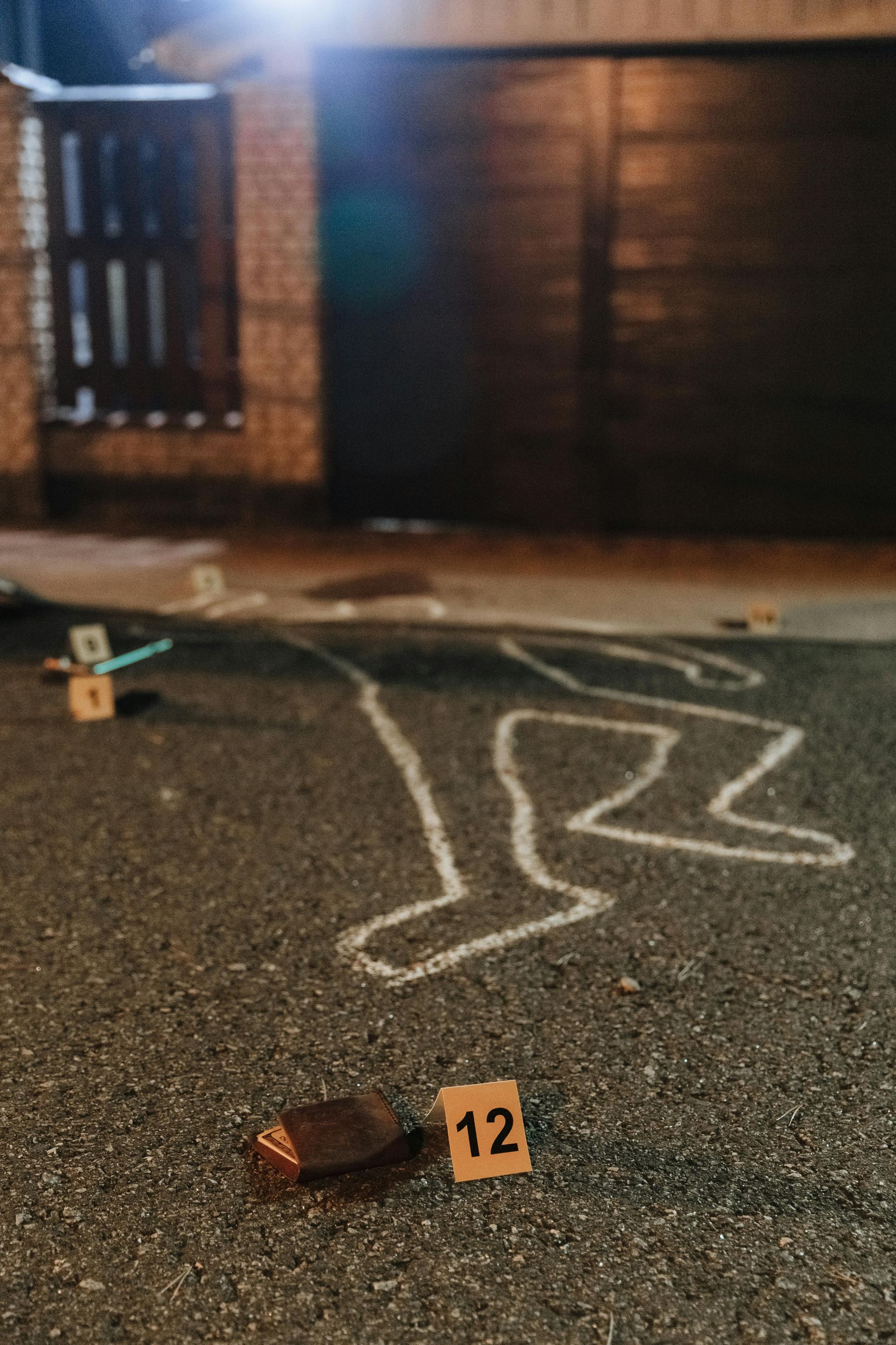 Crime scene: chalk outline of a body on pavement. Evidence markers, garage door, and low lighting.