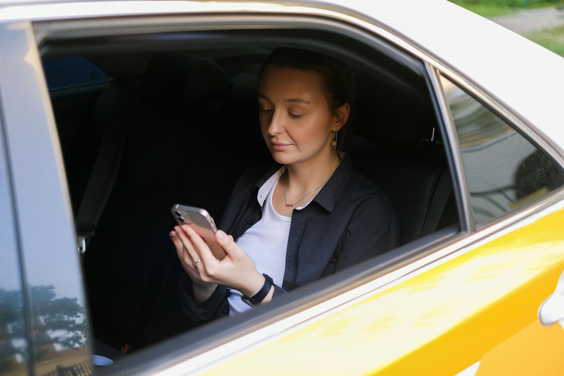 Woman in car, looking at her phone. Black blazer over white shirt. Daytime, outdoors.