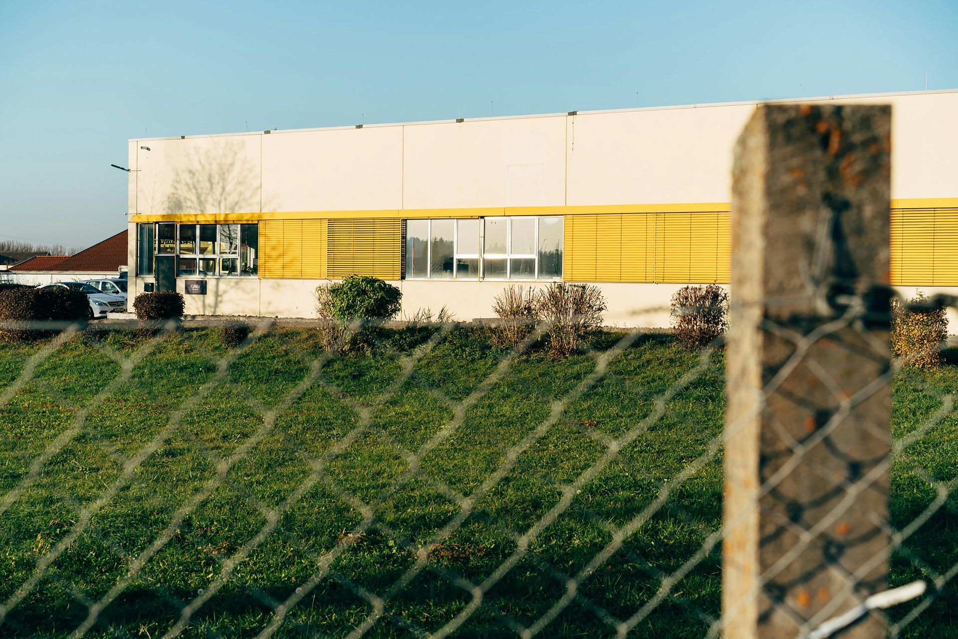 A building with yellow stripes, seen through a chain-link fence and grassy hill under a blue sky.