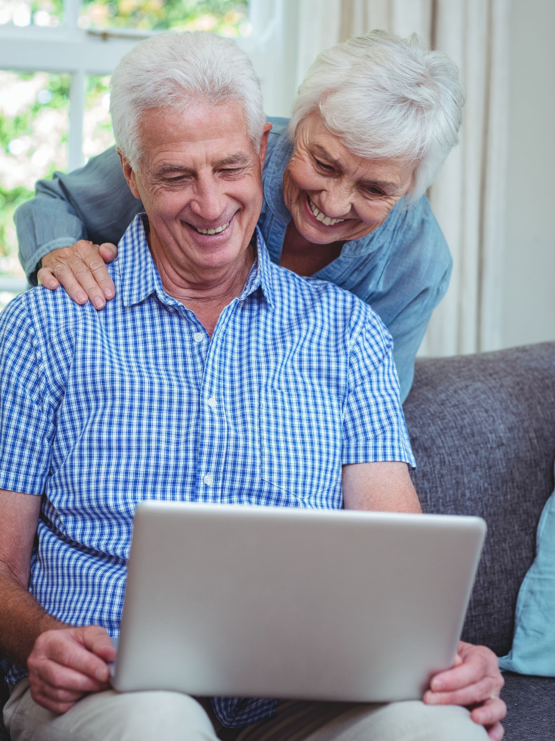 Elderly couple smiles while looking at a laptop on a couch. Woman has arm around man.