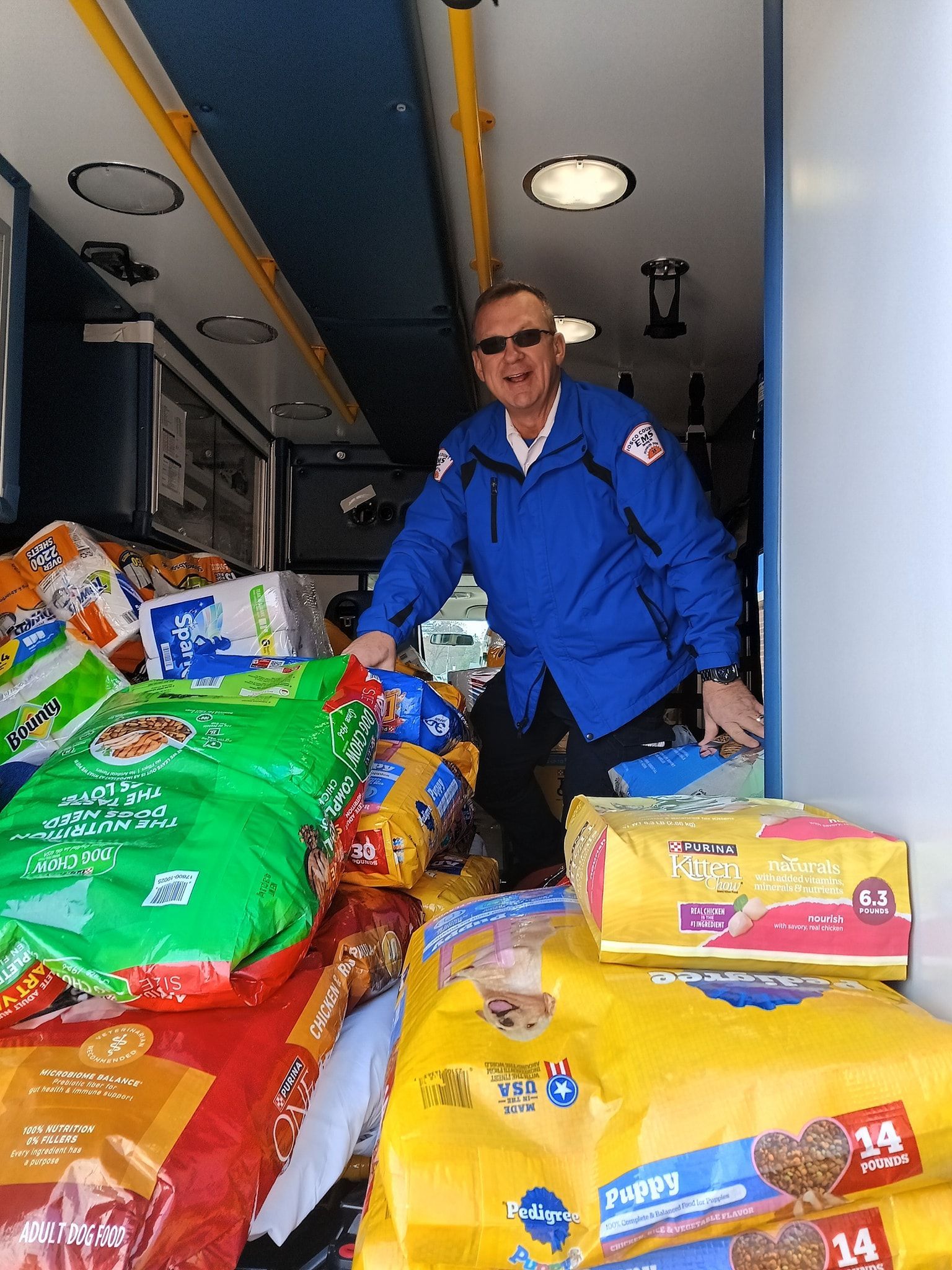 A tabby cat looks up at the camera while standing on a blue and yellow tiled floor near a food bowl.