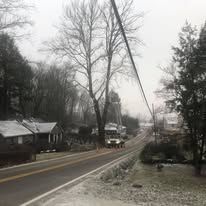 A utility truck on a snow-covered road near a leafless tree, houses and power lines in the background.