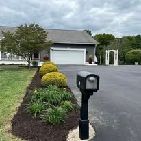 Mailbox on a black post in front of a house, driveway, and landscaping with bushes.