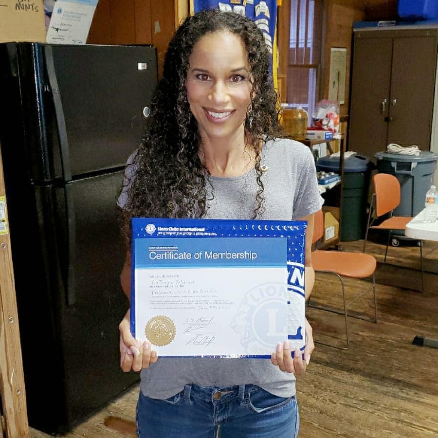 Woman smiles, holding a certificate, near a refrigerator, possibly indoors.