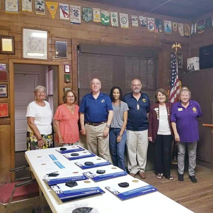 Group of people standing around a table, likely a club meeting. Wooden interior with flags and awards.