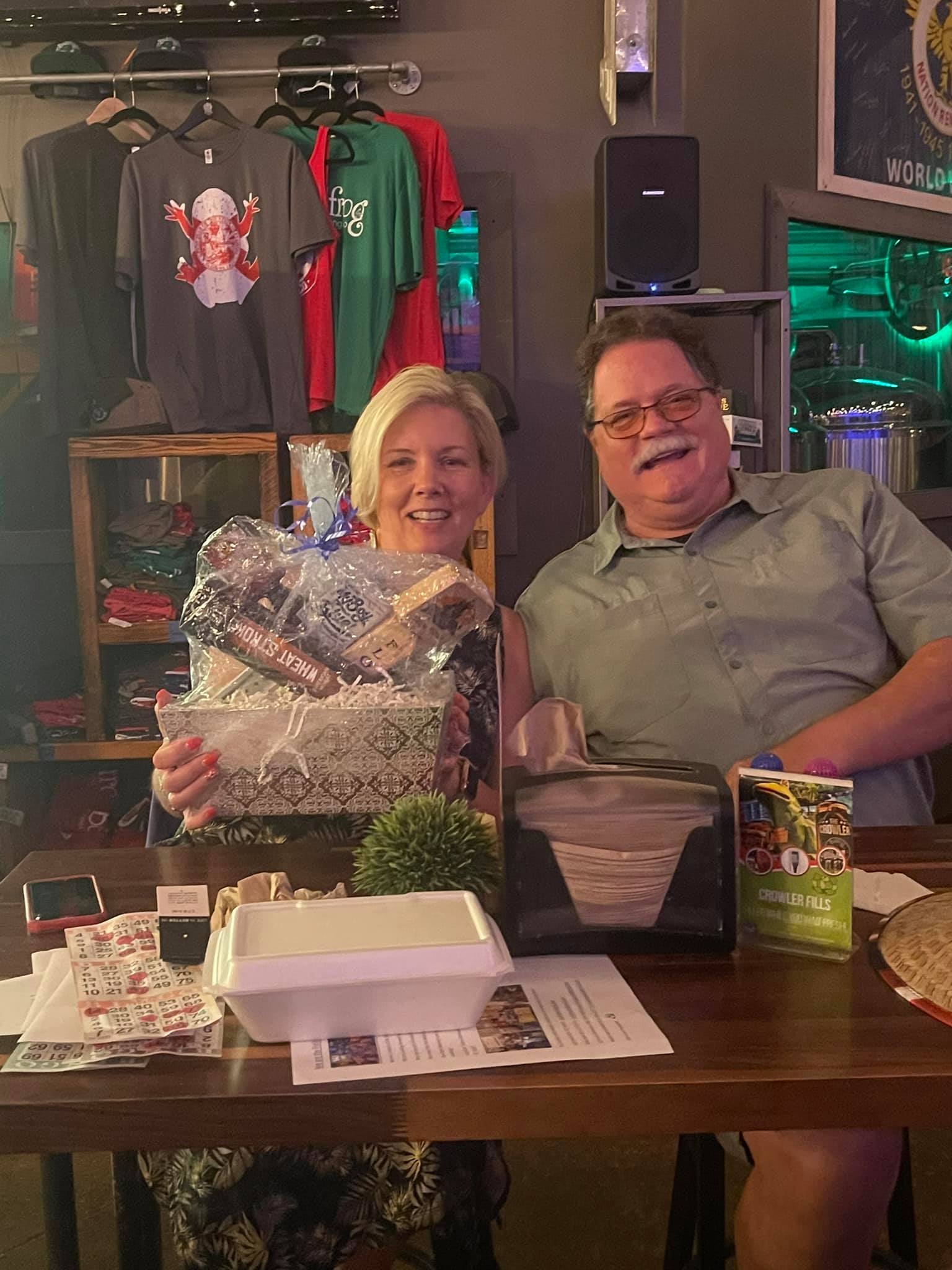 Two people smiling, holding gifts at a bar. Woman in sparkly silver wrap, man in a green shirt.