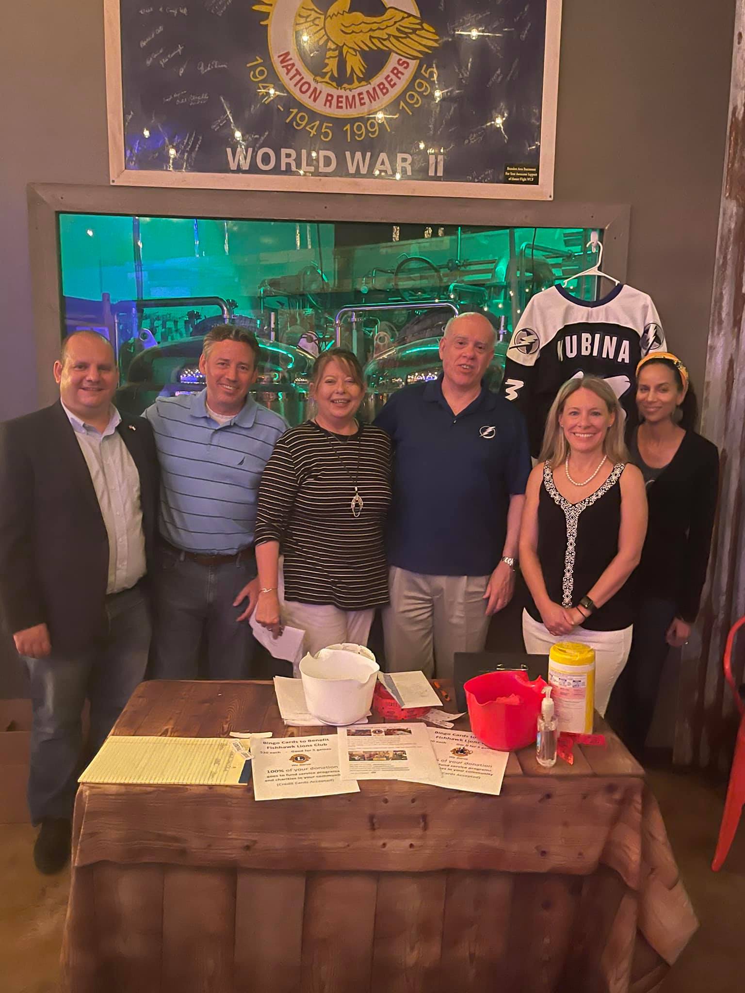 Group of seven people standing behind a wooden table with papers, drinks, and a flag; indoors.