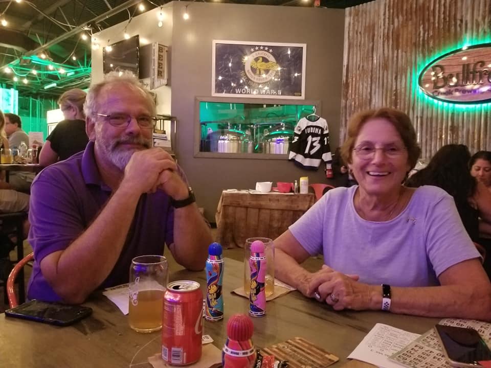 Two people at a table in a bar. Man with a beard smiles, woman smiles. Drinks and decor visible.