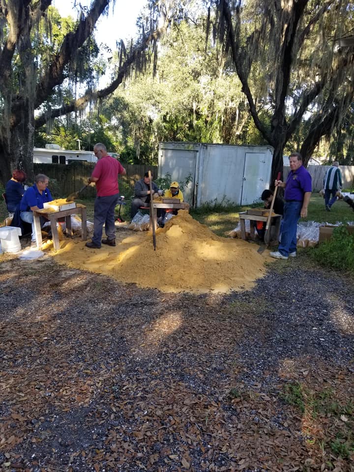 People husking corn outdoors at tables, pile of husks in the center, trees and a shed in the background.