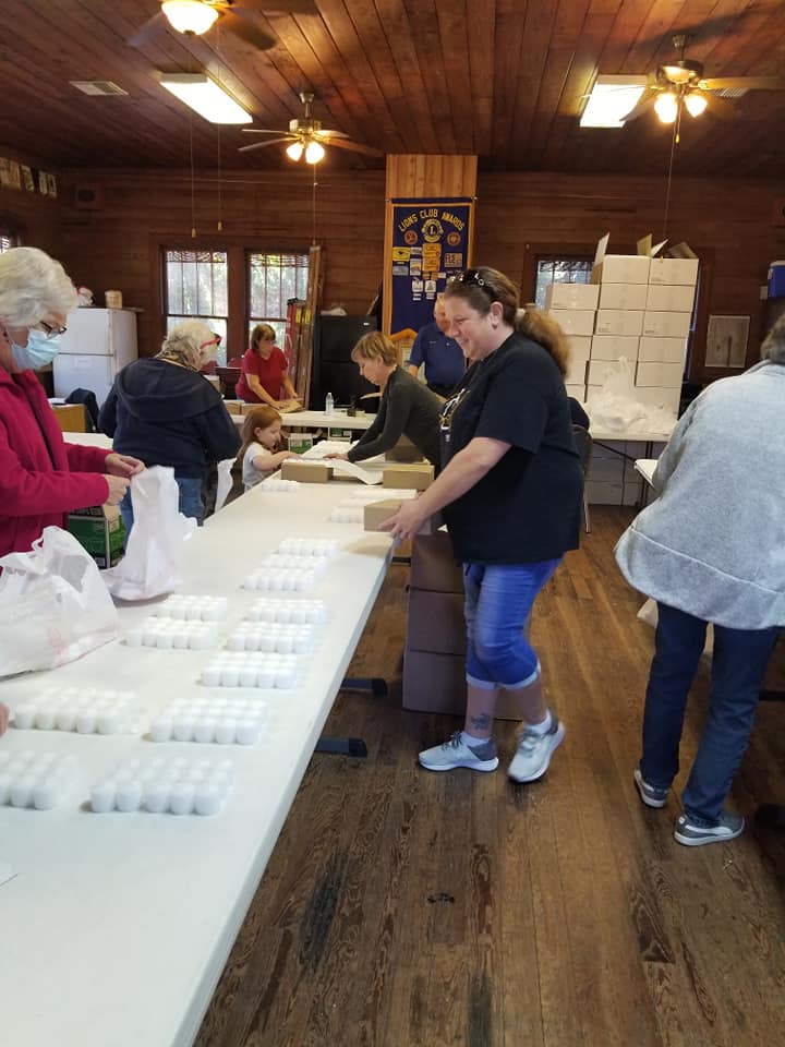 People packing boxes, likely food, in a community hall. Volunteers sort items at long tables, setting up distributions.