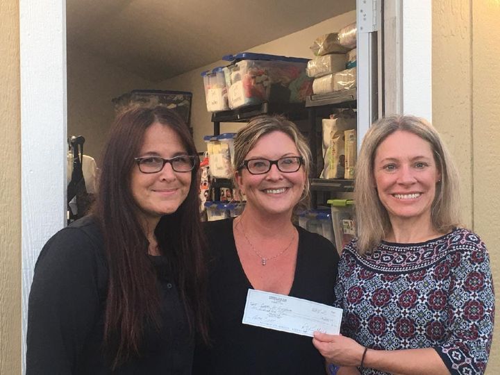 Three women smiling, one holding a check, in front of a food storage area.