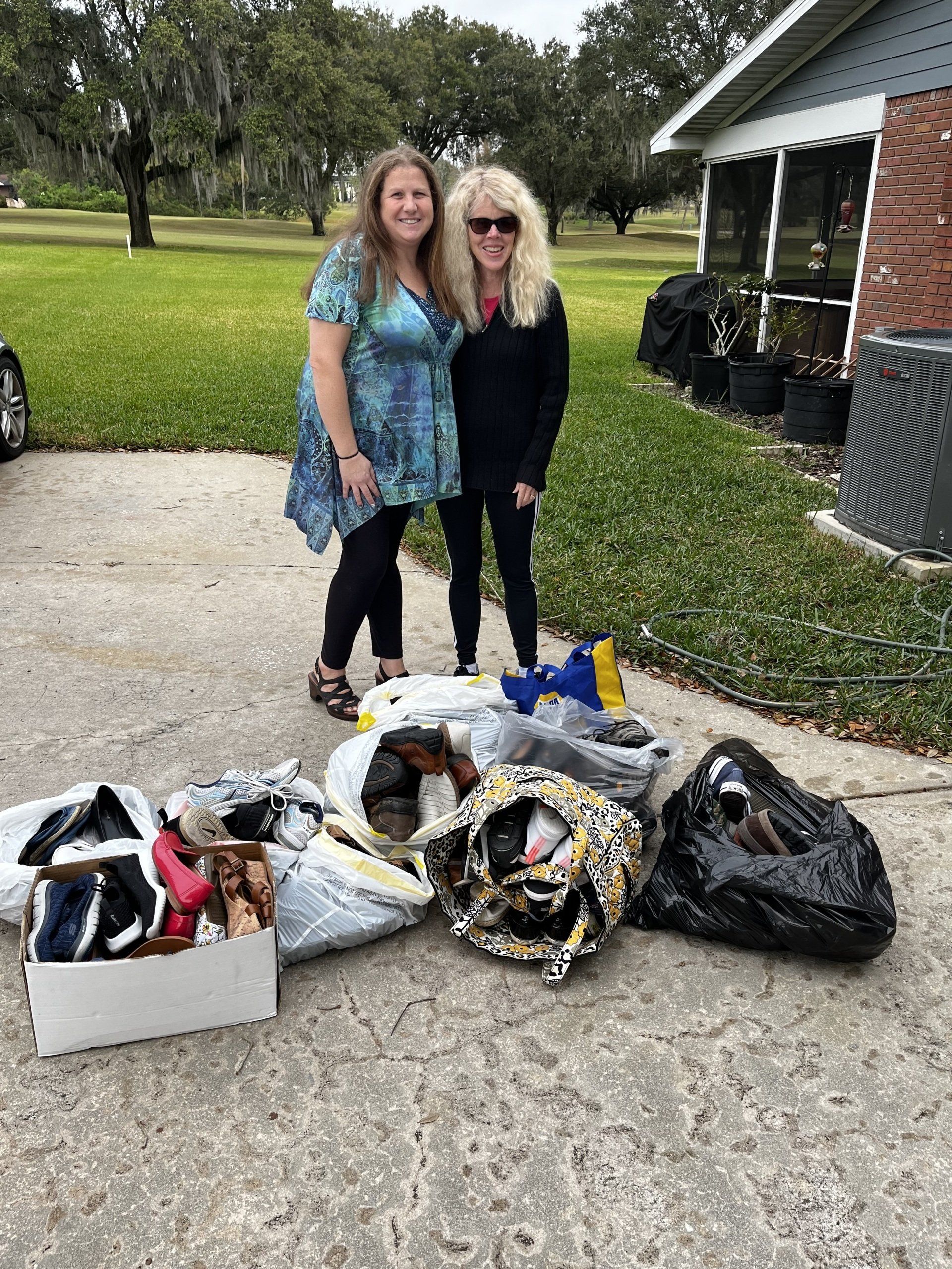 Two women stand with bags of items in front of a house. Bags are grey, black, and patterned.