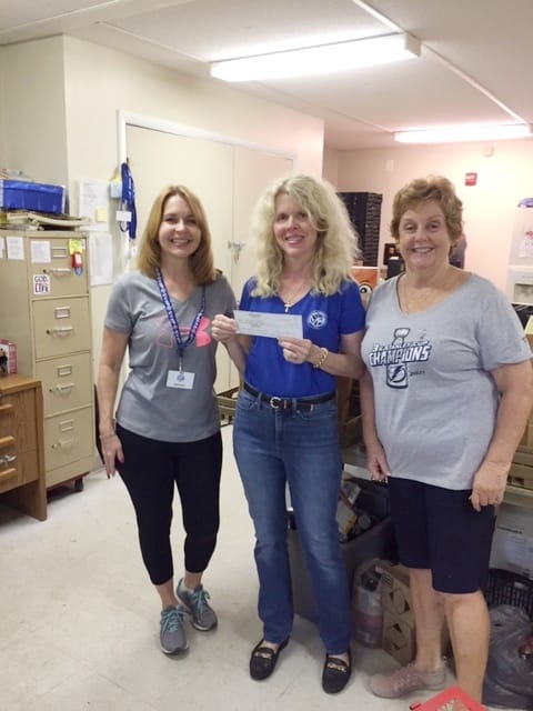 Three women in a room; one in blue holding a check, smiles. Two in grey outfits, one smiles. Office setting.