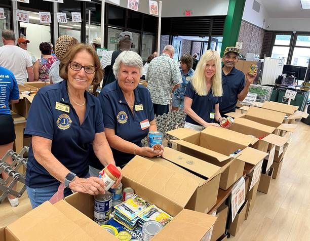 Volunteers in blue shirts packing food boxes in a well-lit room. They smile and sort items like peanut butter.