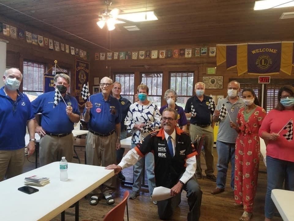 Group of people, some wearing masks, holding checkered flags in a wood-paneled room. Lions Club banner visible.