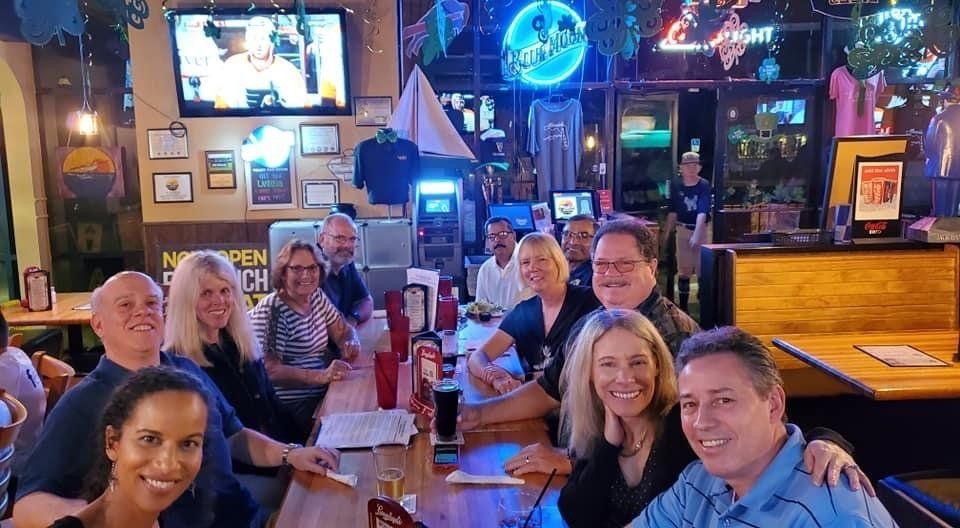 Group of people smiling, gathered at a bar. Wooden tables, drinks, and a TV in the background.