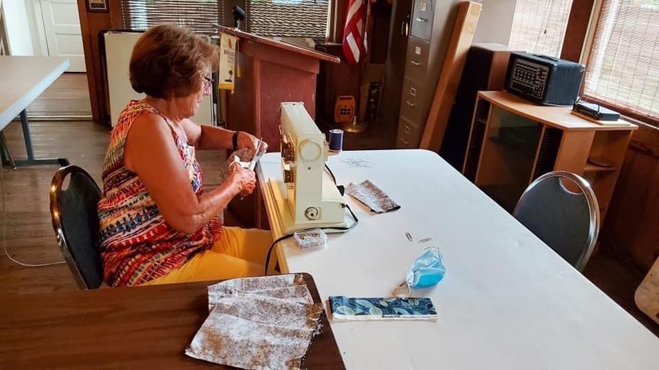 Woman sews at a table indoors; red/orange top, yellow pants; sewing machine; white table, wooden interior.