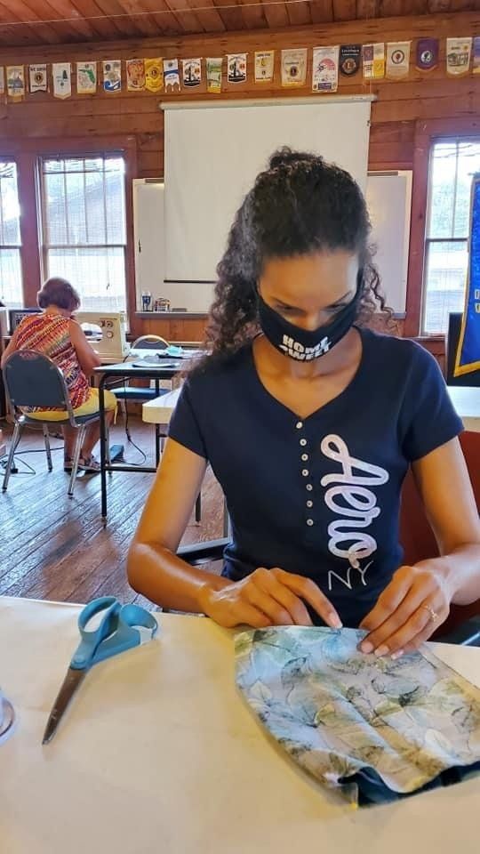 Woman sewing fabric mask in a room. She is wearing a mask and a navy shirt. Blue scissors on the table.