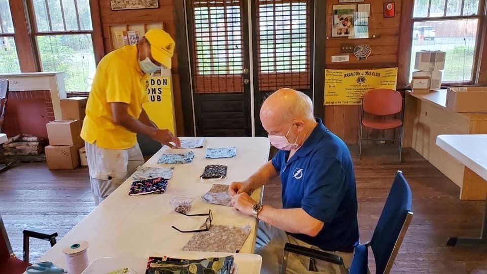 Two men in masks arranging fabric face masks on a table inside a room.