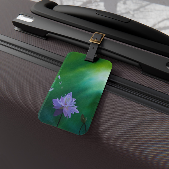 green luggage tag featuring a purple lotus flower with mini butterflies flitting by fixed to a gray suitcase by a black leather strap 