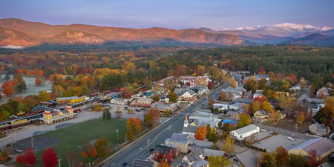 Aerial view of a town nestled in colorful autumn foliage with mountains in the background.